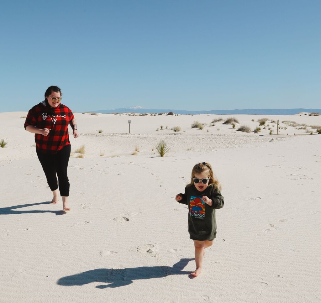 Editor, beta reader, and copywriter Kyare Betzing chasing her daughter in the White Sands of New Mexico with snow-capped mountains and blue skies in the background.